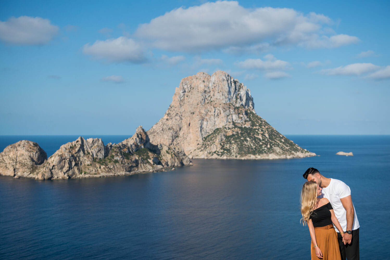 couple standing in the sun on a clifftop with an island as the backdrop in the sea
