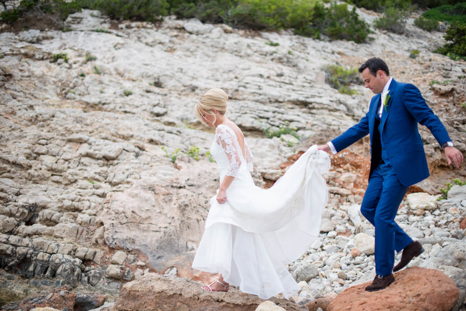 Groom holding up his bride's veil while walking across the rocks at their Ibiza wedding