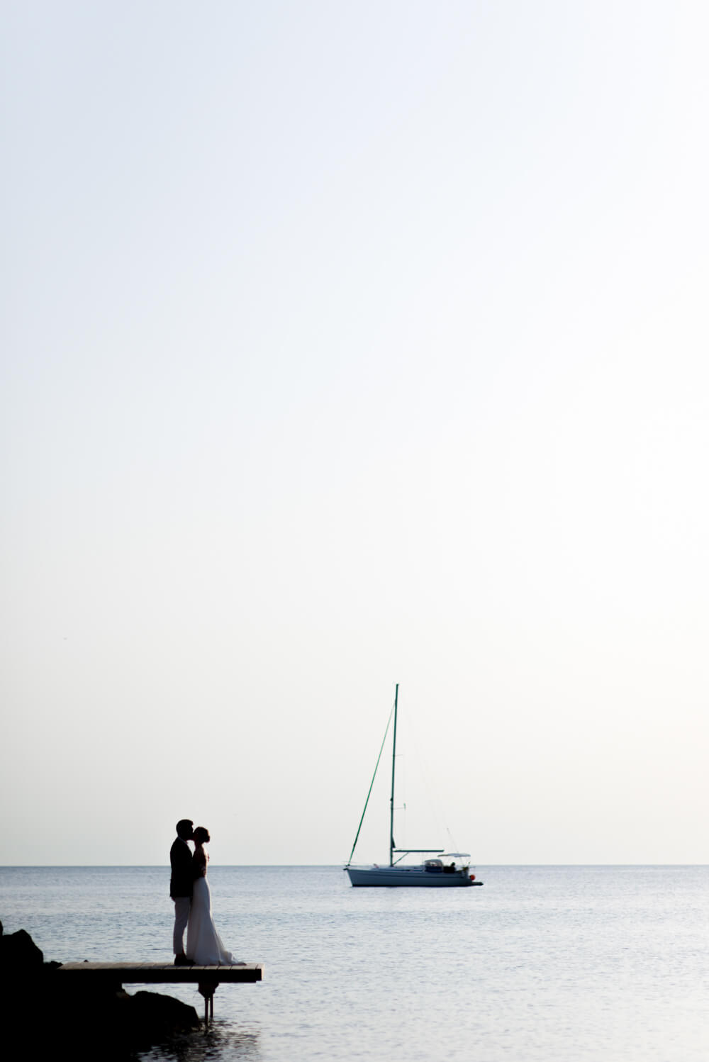 bride and groom kissing on a pier with a sailing boat in the background at their wedding in Ibiza