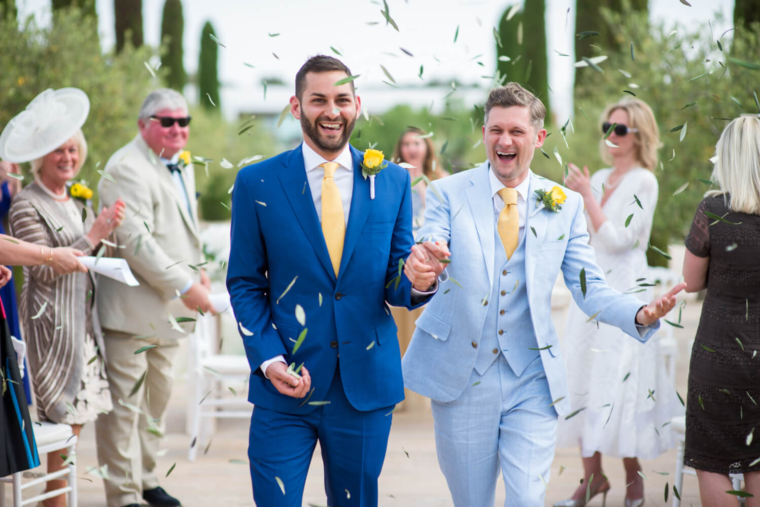 Two grooms walking out of their ceremony with confetti of olive leafs at Ca Na Xica in Ibiza
