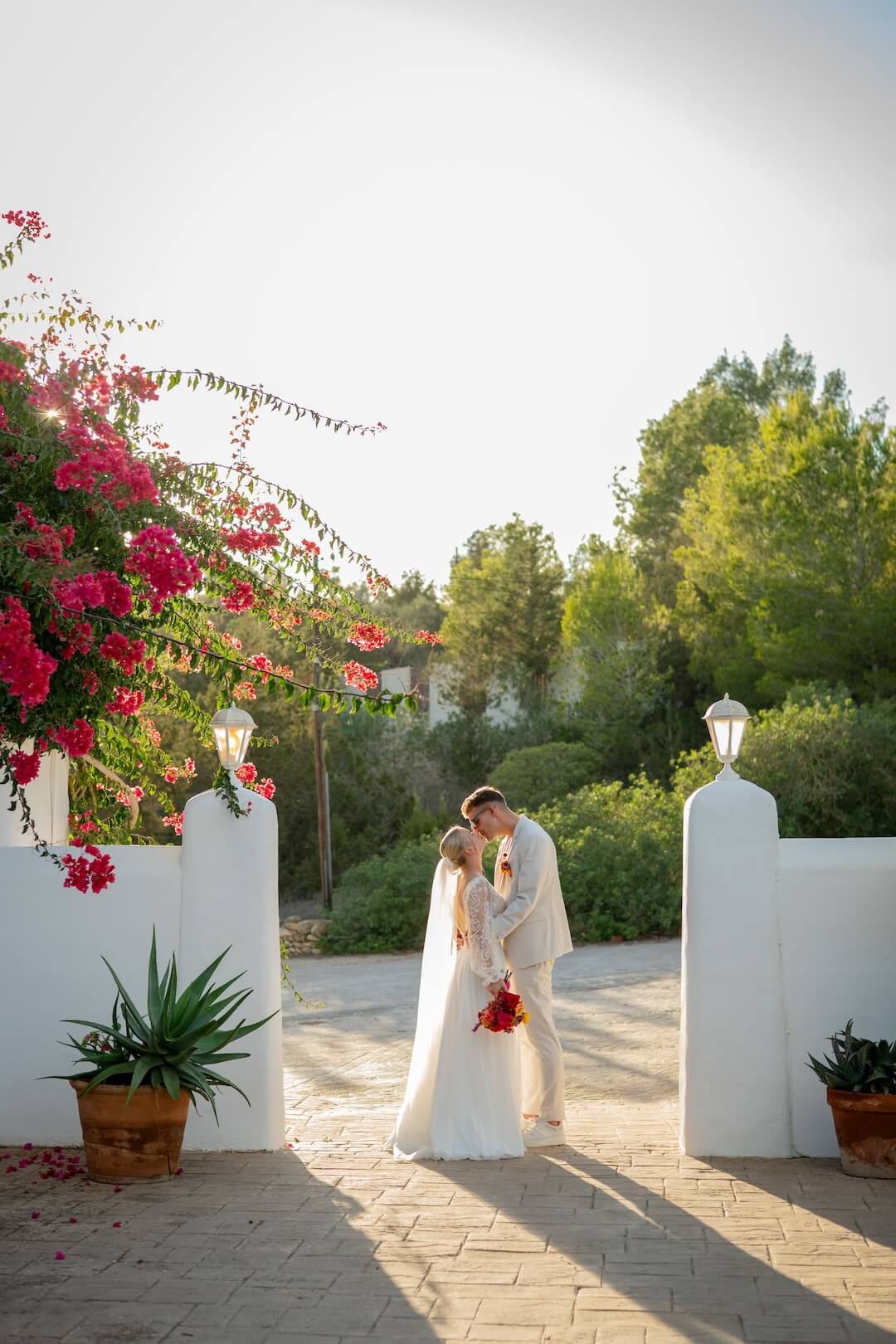newly weds, husband and wife stand together under the golden hour light during sunset in ibiza after their wedding ceremony. white dress on, kissing in between white walls at Can Domingo
