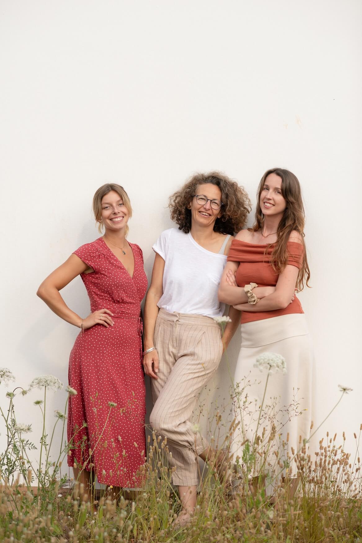 three women standing together posing for photo against white wall in ibiza. wearing neutral and red tones with grass below covering their feet